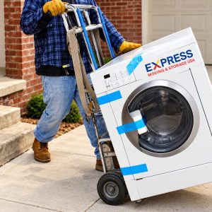 A worker wearing gloves and boots uses a hand truck to move a front-loading washing machine labeled EXPRESS MOVING & STORAGE outside a brick-front home.